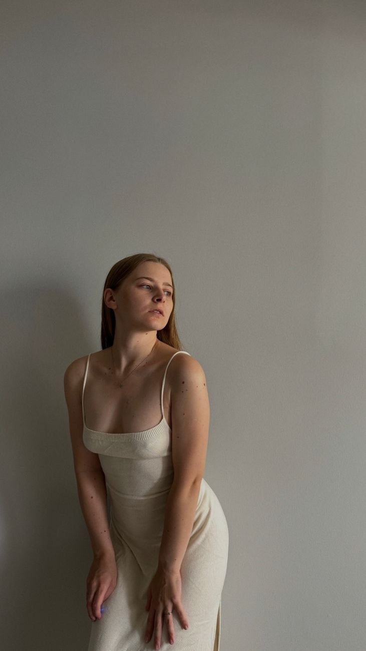 A woman in a cream dress poses elegantly against a minimalist background.