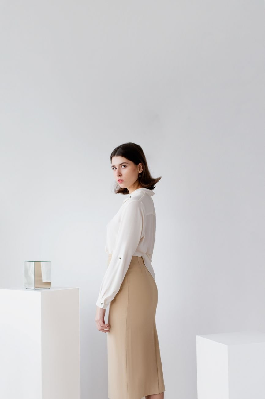A woman poses indoors in a minimalist setting wearing a white blouse and beige skirt.